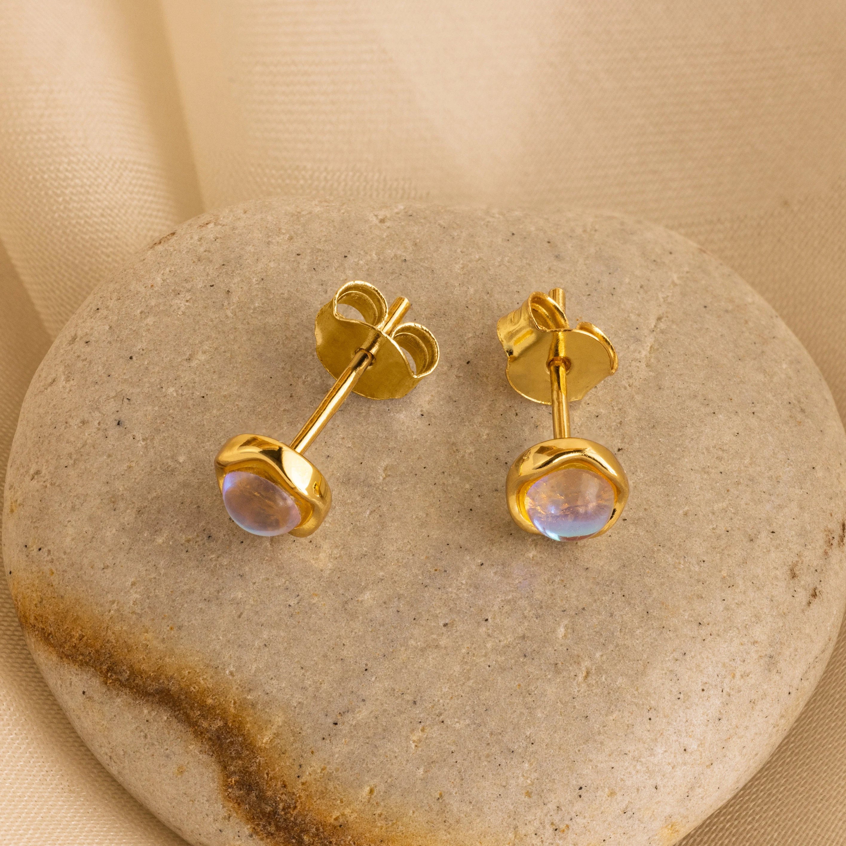 A pair of gold-colored moonstone stud earrings placed on a textured stone surface.